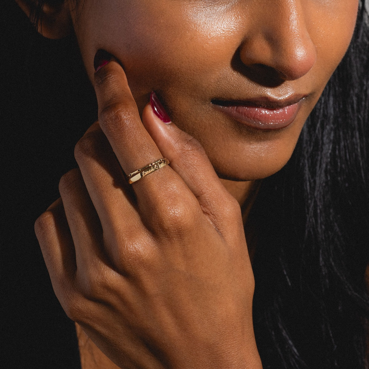 A person with medium brown skin and long dark hair touches their face, showcasing the Solid Gold Tiny Fuck Birthstone Stacking Ring Set. Their glossy lips and dark red nails stand out against a dark background.