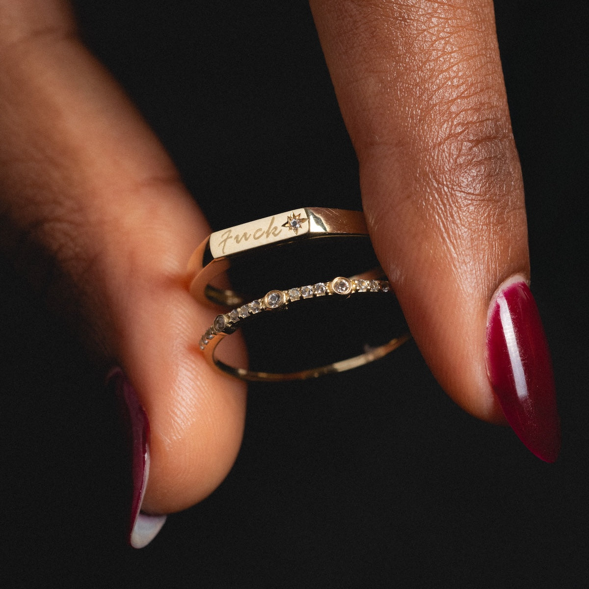 A hand with maroon-painted nails holds the Solid Gold Tiny Fuck Sapphire Stacking Ring Set—one ring engraved with fuck and a small star, and the other adorned with a row of tiny sapphires.