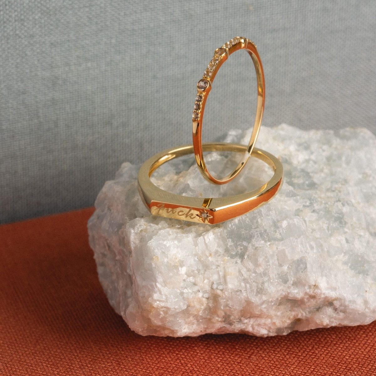 Two solid gold stacking rings rest on a rough white stone. One is thin with small sapphires, and the other is plain with fuck engraved. The background is gray and the surface is rust-colored fabric. Product: Solid Gold Tiny Fuck Sapphire Stacking Ring Set.
