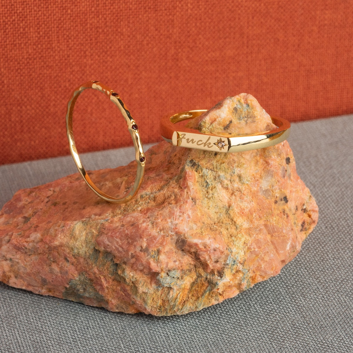 The Solid Gold Tiny Fuck Birthstone Stacking Ring Set features two gold rings on a peach stone: one slim with tiny bumps, the other thicker with fuck and a star engraving. Background fabrics are orange and gray.