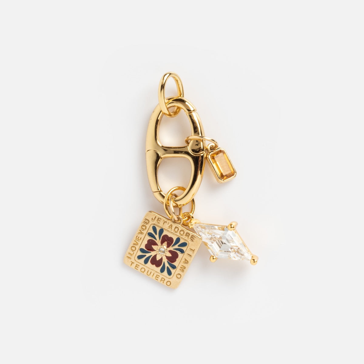 The Charm Clip is a gold vermeil piece with three pendants: a small gemstone, a diamond-shaped clear gem, and a floral square tile inscribed with “Te Quiero” and “Amore.” Ideal as a bracelet charm or unique accessory clip.