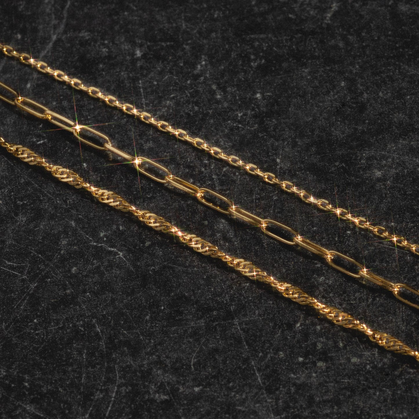 The Oval Link Chain, a gold-plated necklace, is arranged diagonally on a dark textured surface, with light reflecting off the metal for sparkling highlights—a true charm collection.