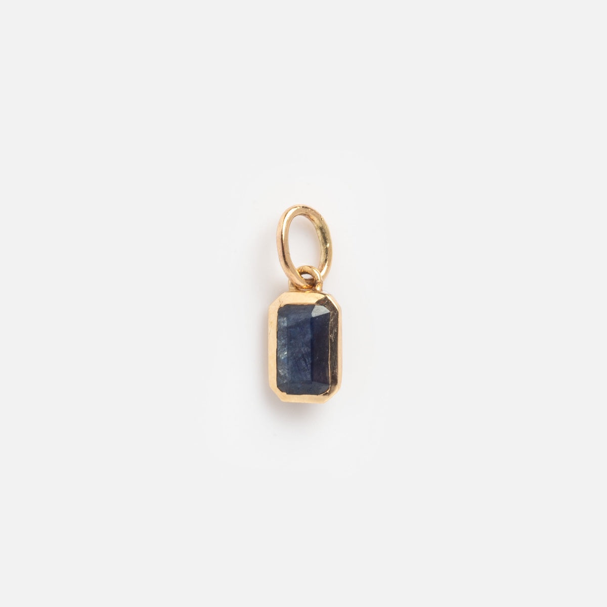 The Solid Gold Birthstone Baguette Charm September Sample features a small, rectangular dark blue gemstone with faceted edges, set in a gold frame and attached to a simple gold loop, shown against a plain white background.