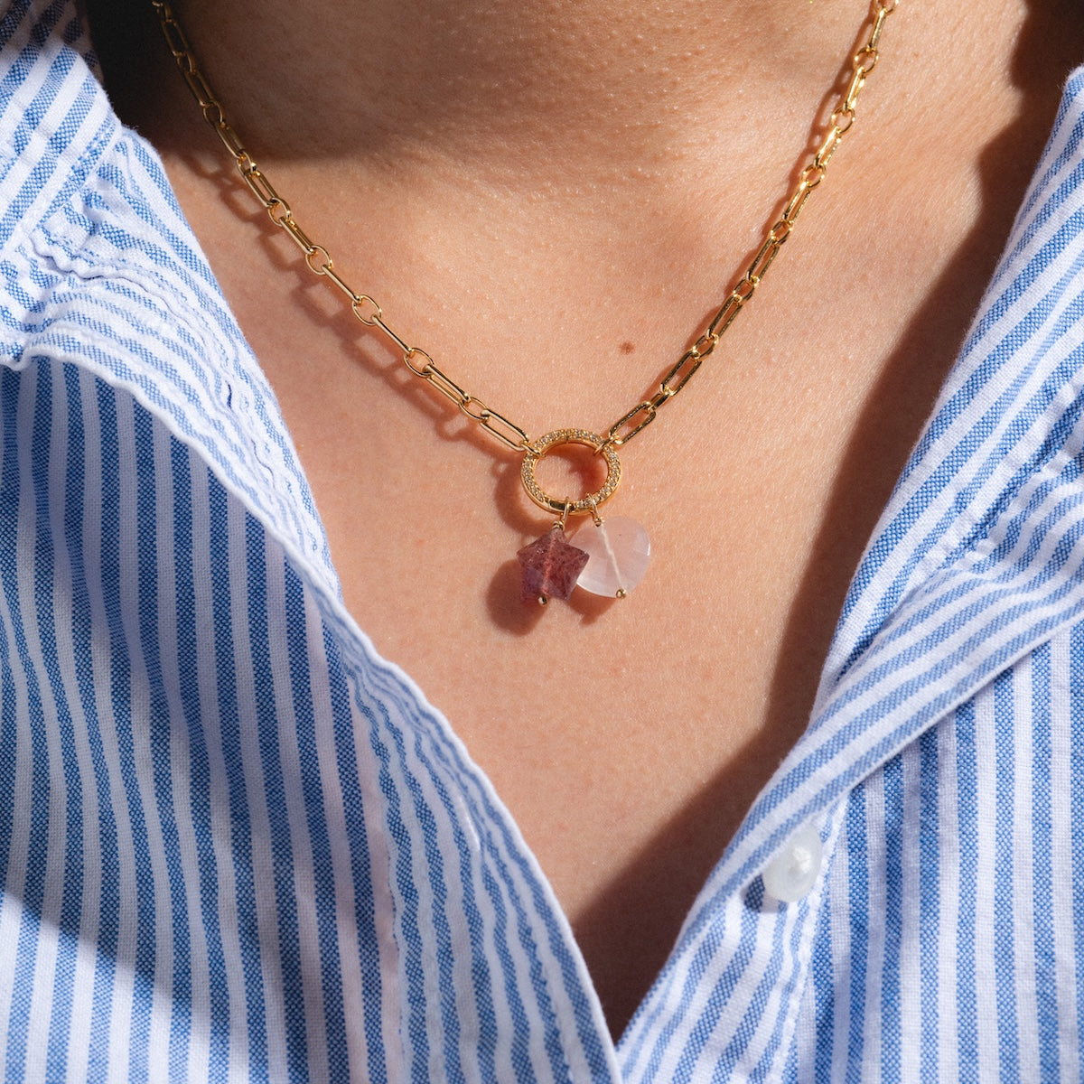 A close-up of a person wearing the Devi Arts Collective x Local Eclectic Carved Crystal Charms gold chain necklace with pink and purple gemstone pendants, styled over a blue and white striped button-up shirt—a chic semi-precious stone jewelry look.