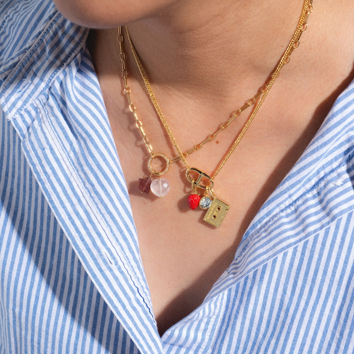 A person in a blue and white striped shirt wears two layered gold necklaces with charms, including the Devi Arts Collective x Local Eclectic Carved Crystal Charms, a heart, moonstone charm, red beaded berry, and rectangular pendant.