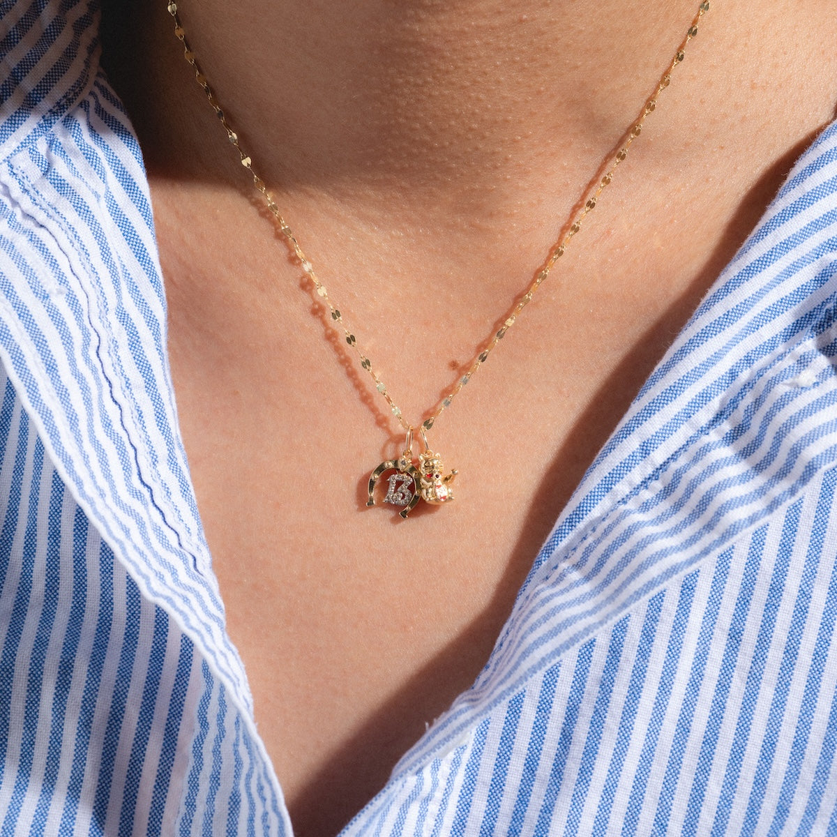 A close-up of a person wearing the Solid Gold Lucky Charm necklace with small charms, paired with a blue and white striped collared shirt.