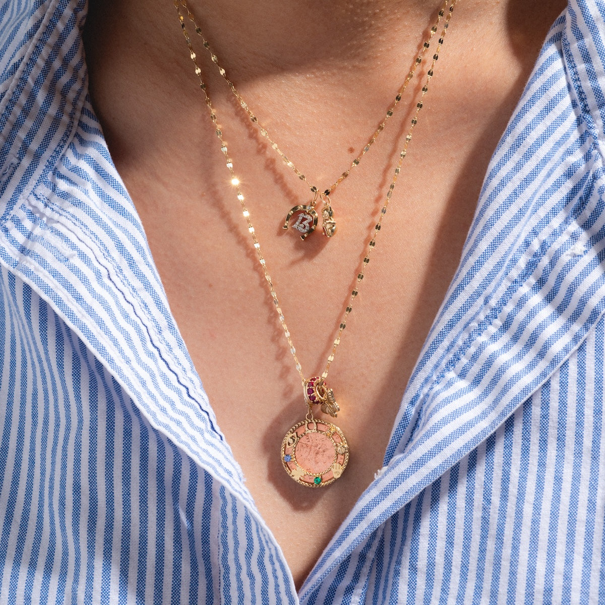 A person wears a blue and white striped shirt with two layered gold necklaces—one features a circular pendant with pink and green accents, and the other displays the Solid Gold Lucky Charm as a small, intricate detail.