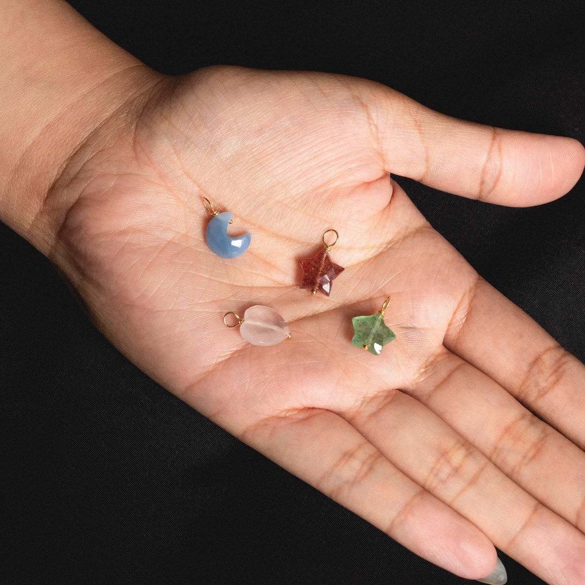A hand displays four Devi Arts Collective x Local Eclectic carved crystal charms—crescent moon, star, teardrop, and leaf—against a black background, ideal for fans of semi-precious stone jewelry.