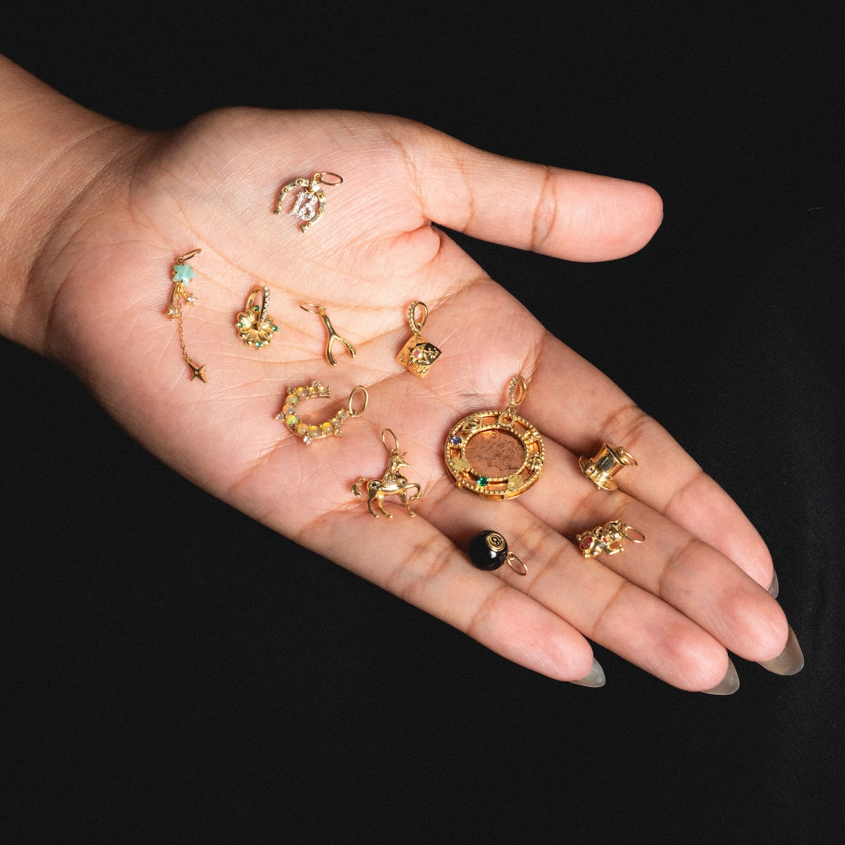 A hand with long nails holds several Solid Gold Lucky Charms, each featuring unique designs and colorful gemstones, set against a black background.