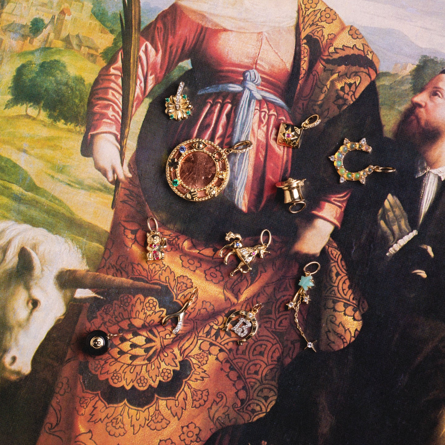 The Solid Gold Lucky Charm, adorned with colorful gemstones, is displayed on patterned fabric with a classical painting of a woman in historical attire and a man in the background, alongside part of a white animal.