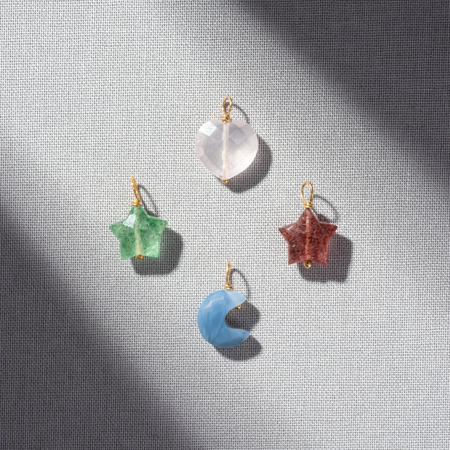Four Devi Arts Collective x Local Eclectic Carved Crystal Charms with gold loops—clear heart, green star, red star, and blue crescent moon—rest on gray textured fabric, evoking semi precious stone jewelry.