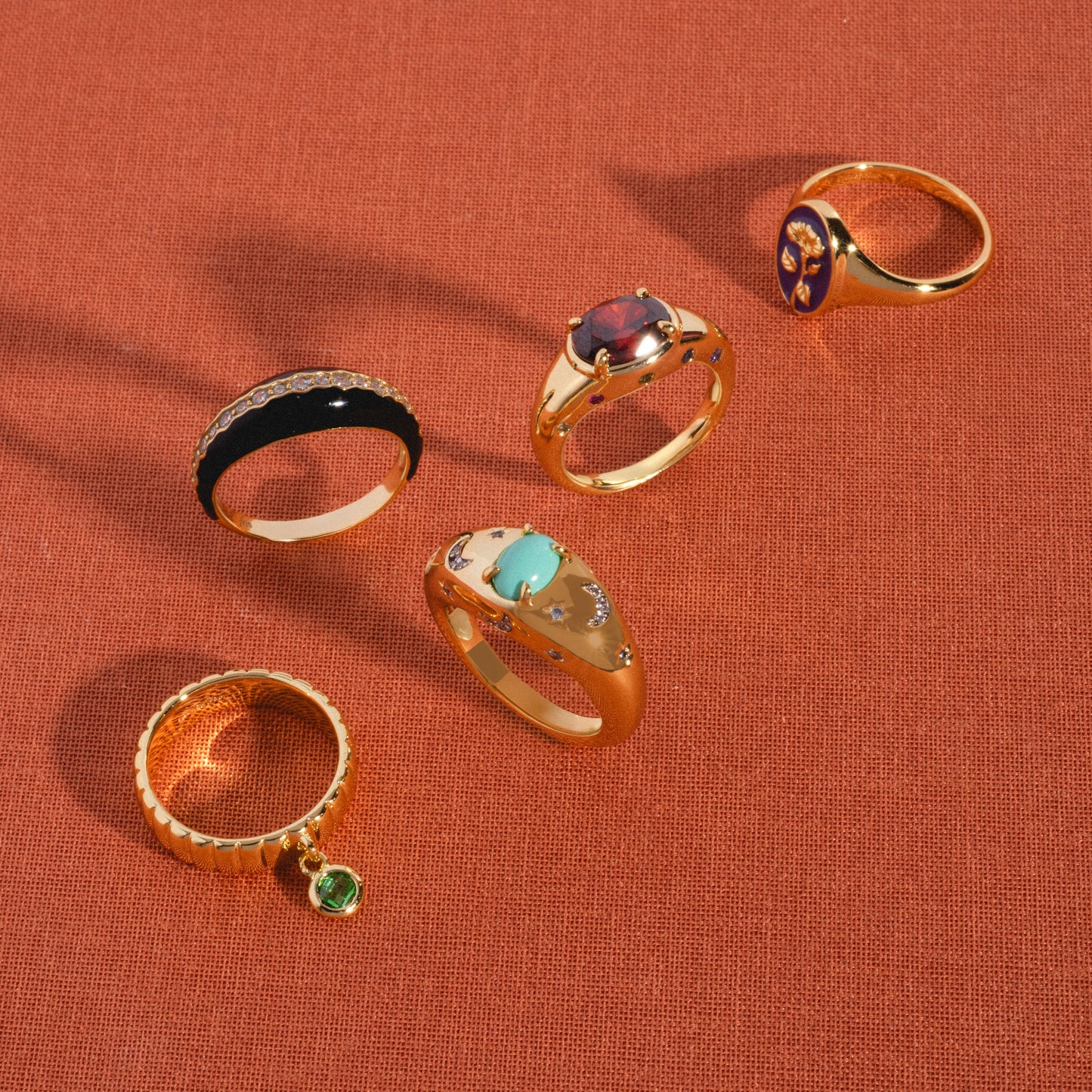 Five gold rings, including the Free Spirit Black Enamel Ring, feature colorful gemstones and unique engravings. Arranged on orange textured fabric, each displays charming details like colored stones or intricate patterns.