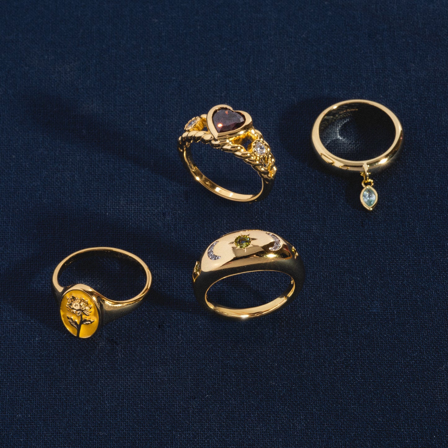 Four 14k gold plated rings on dark blue fabric: the Bold Heart Statement Ring with a heart-shaped stone and diamonds, a ring with a dangling teardrop gem, one with a raised tree design, and one featuring a small green stone in the center.