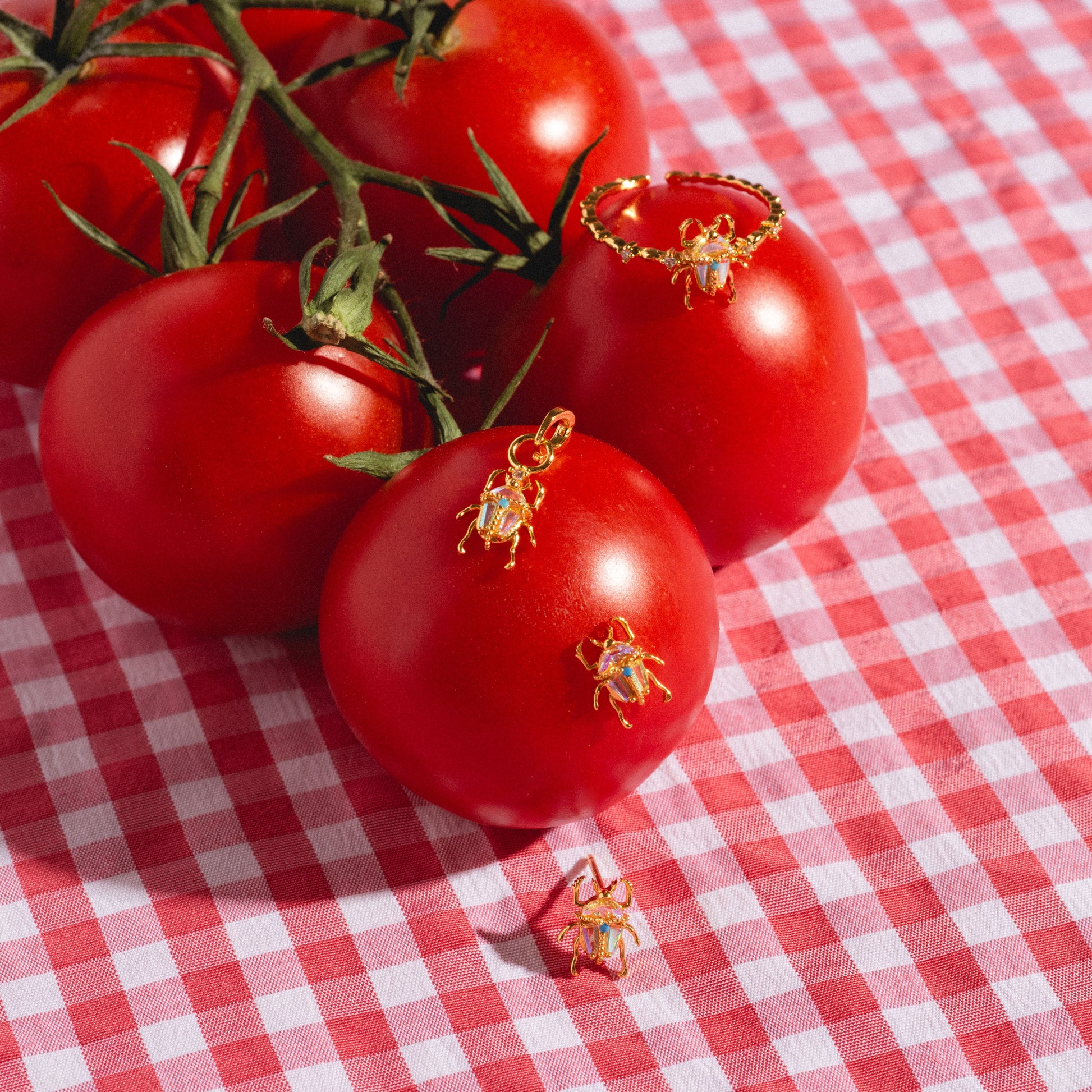 Beetle earrings studs, ring, and charm laid on tomatoes on top of red gingham table cover