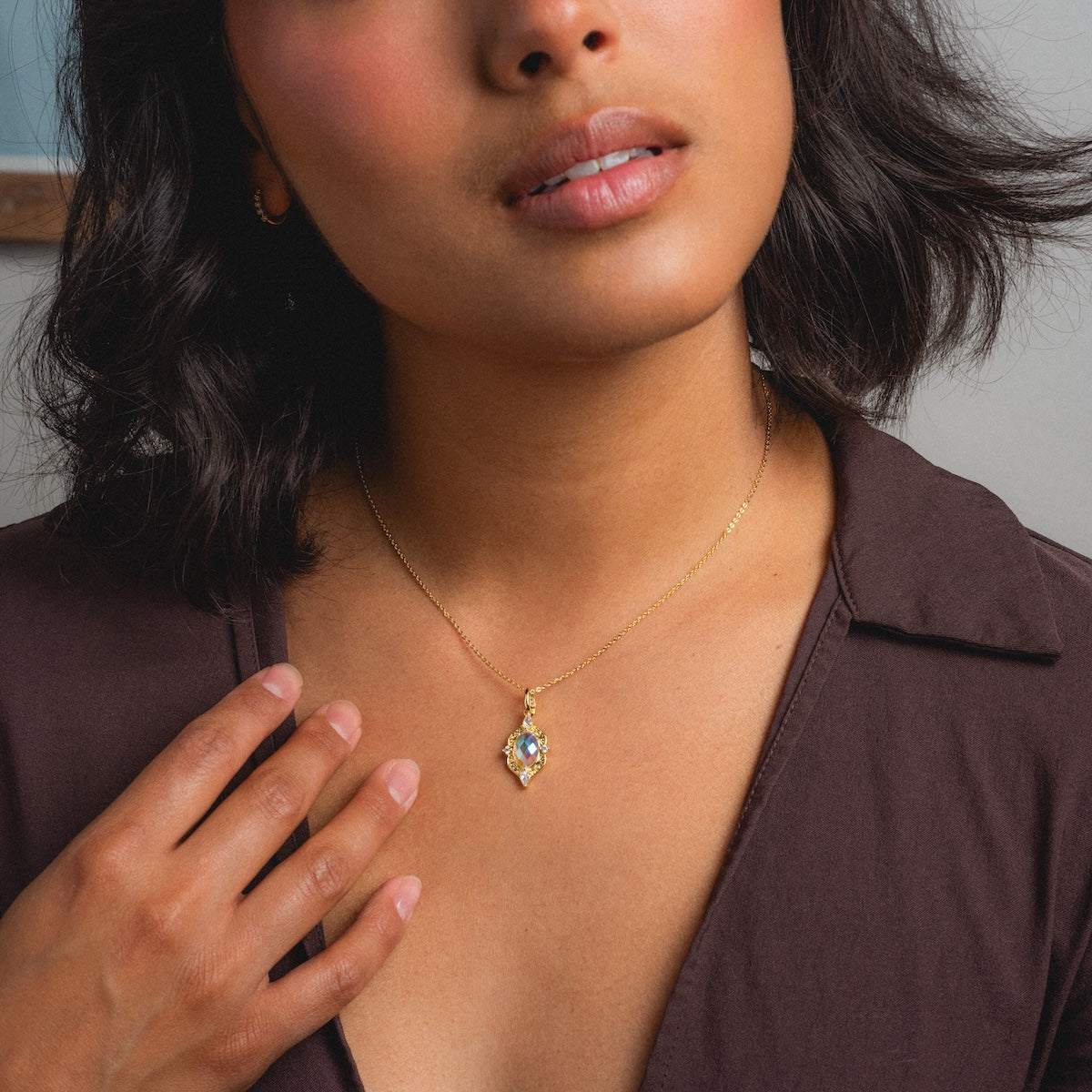 A woman with short dark hair wears a brown blouse and the Lady Luxe Angel Aura Quartz Pendant Necklace, gently touching her collarbone with one hand.