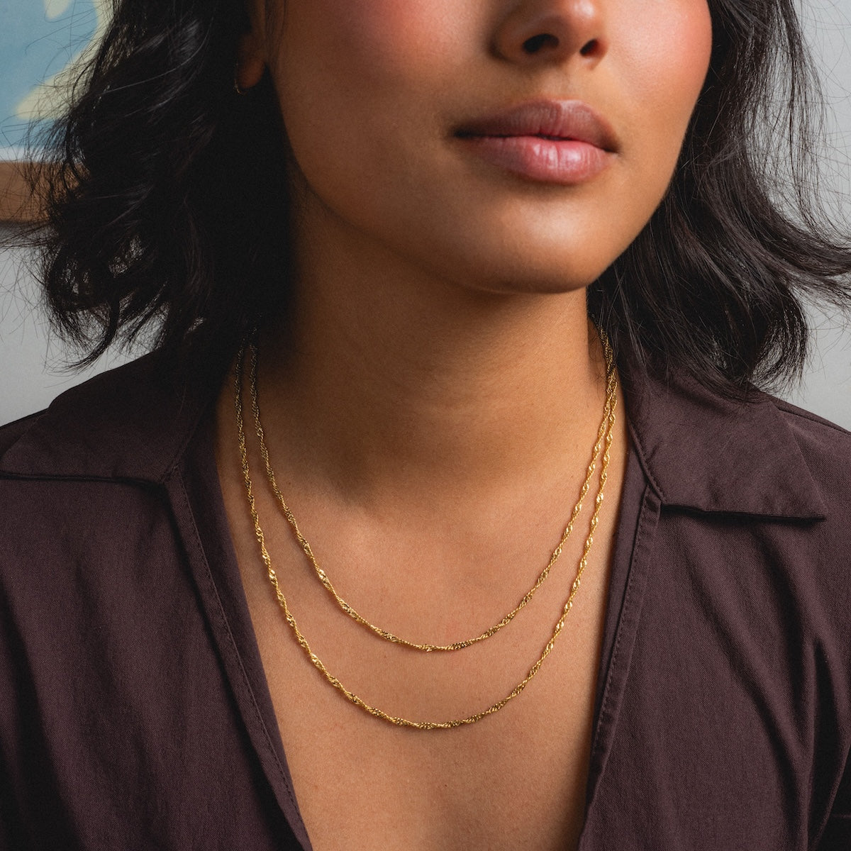 A woman with medium skin tone and wavy dark hair wears the Twisted Rope Chain gold plated layered necklace with a brown button-up shirt; only her lower face and neck are visible.