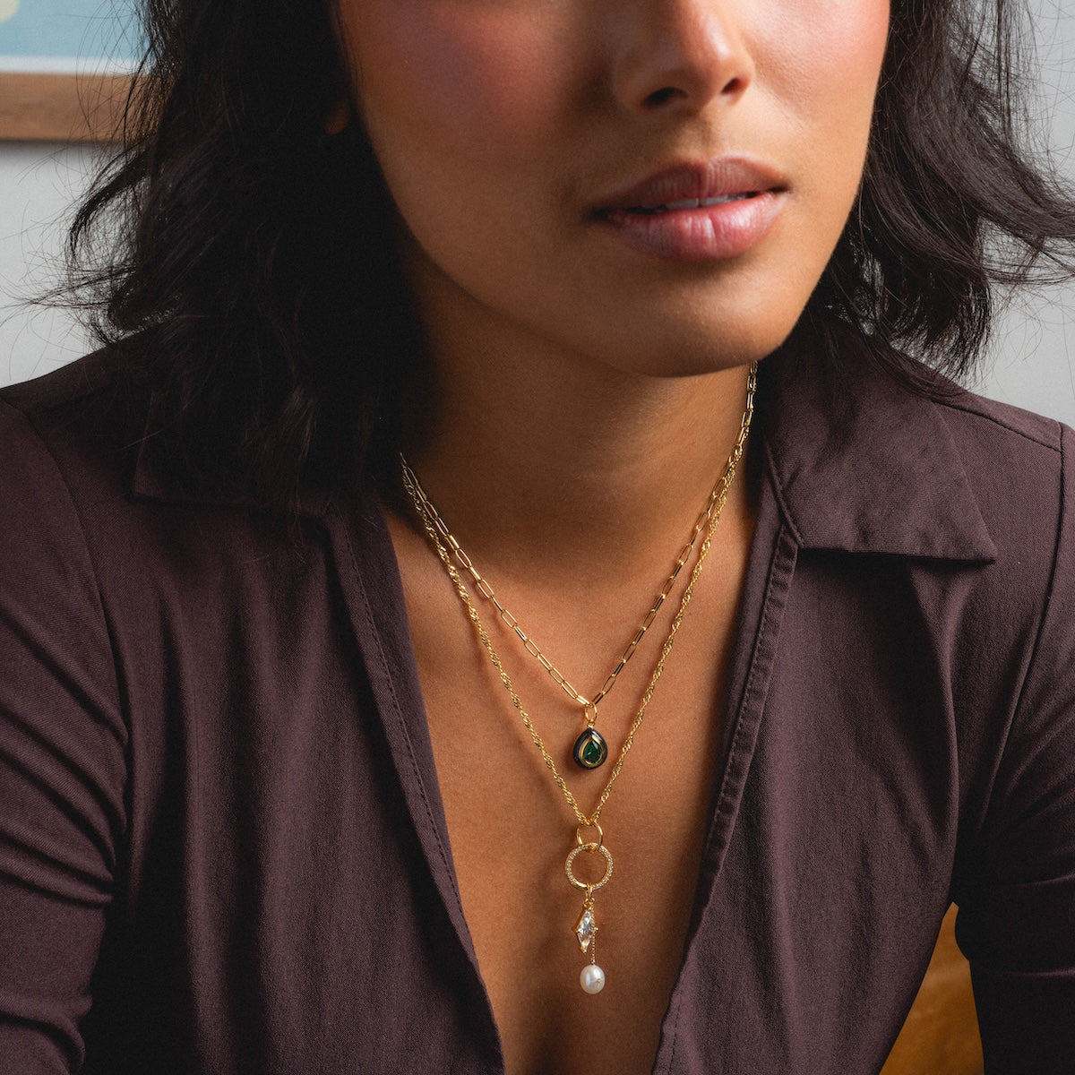A woman, shown from her upper chest with only the lower face visible, wears a brown blouse and two yellow gold plated Charms by Jurate JLA necklaces: one with a green gemstone pendant, the other featuring a pearl and circular charm.