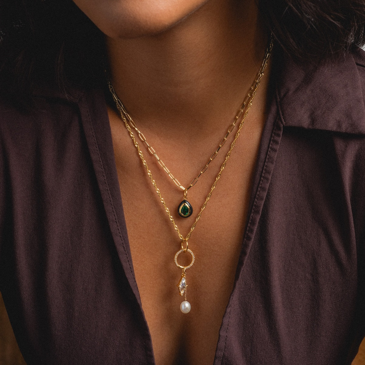 Wearing a dark purple shirt and a gold-plated Twisted Rope Chain necklace with a black teardrop pendant, layered with another chain featuring a circular, crystal-adorned pendant and dangling pearl.