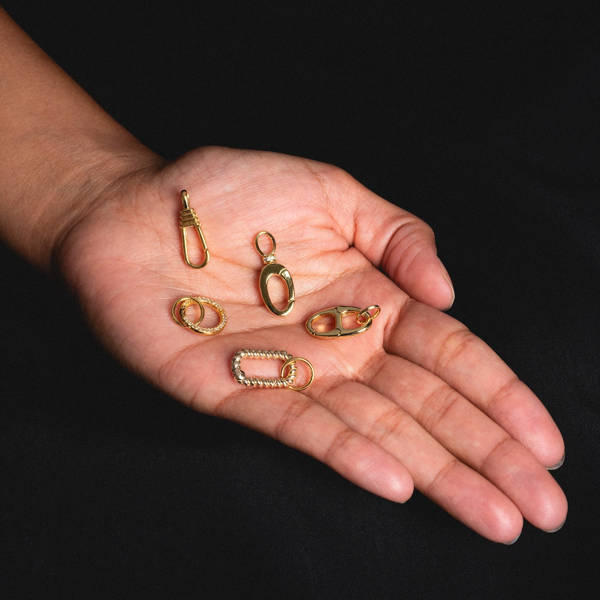 A hand with an open palm displays five Charm Clip gold vermeil findings in various shapes, ideal for bracelet charms, against a black background.