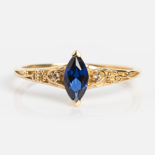 The Solid Gold 2025 September Capsule Classic Blue Sapphire Statement Ring features an ornate engraved band and a marquise-cut lab-created blue sapphire centerpiece, displayed against a plain white background.