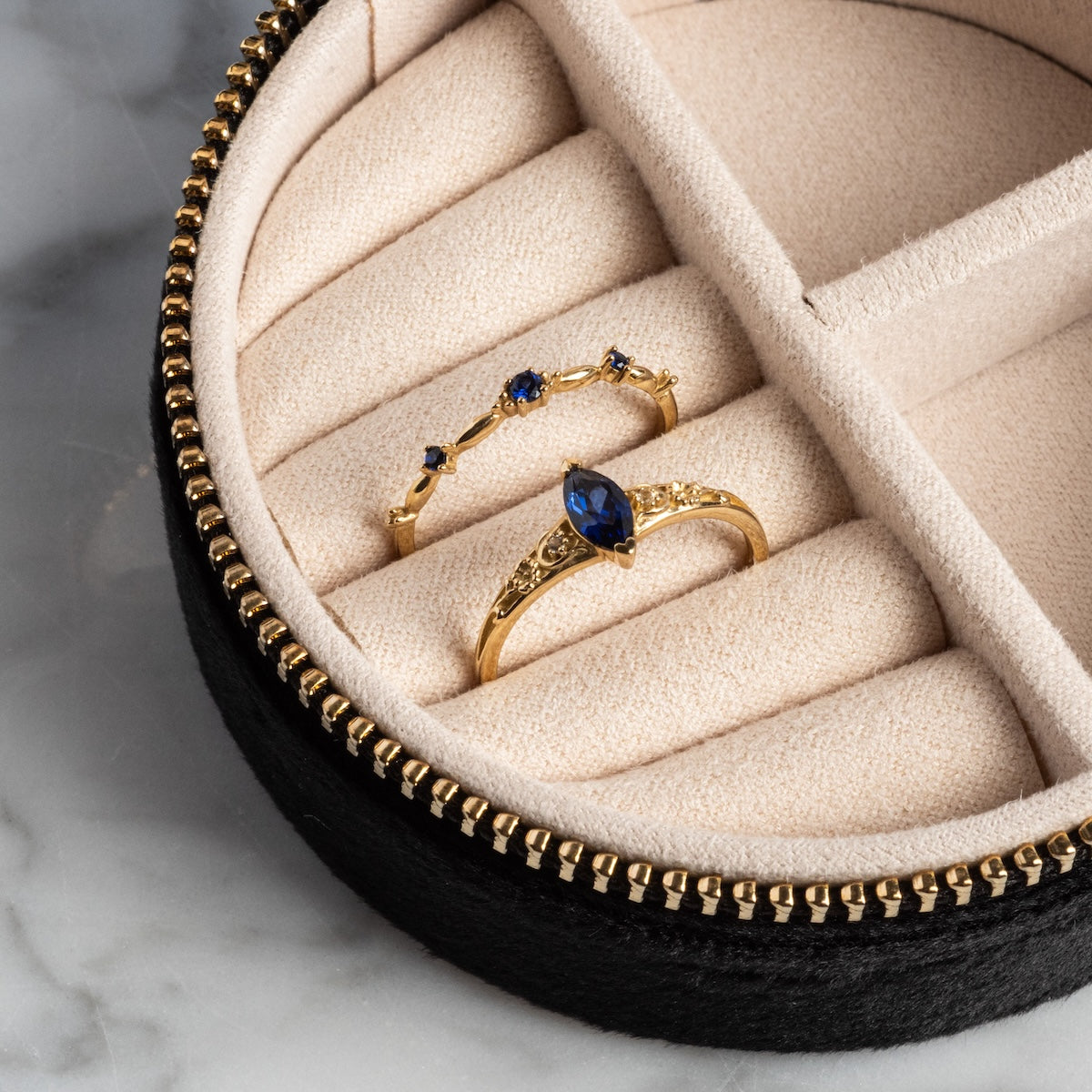 The Solid Gold 2025 September Capsule Blue Sapphire Ring Set is displayed in a beige, velvet-lined jewelry box with a black exterior and gold zipper, placed on marble. Comes with a free gift.