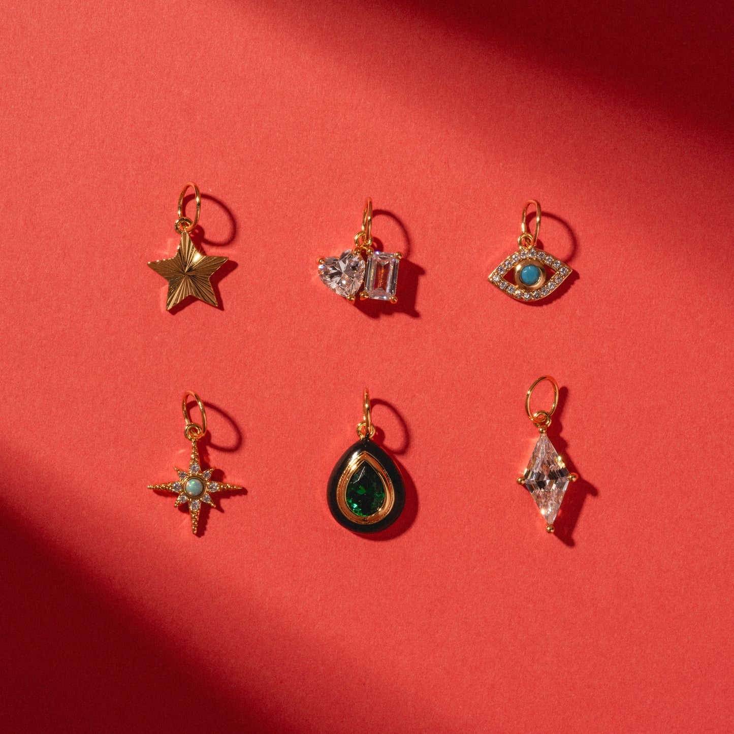Six Charms by Jurate JLA, yellow gold plated with gemstone and simulated diamond accents, are arranged on a red background in star, heart, eye, starburst, teardrop, and diamond shapes.