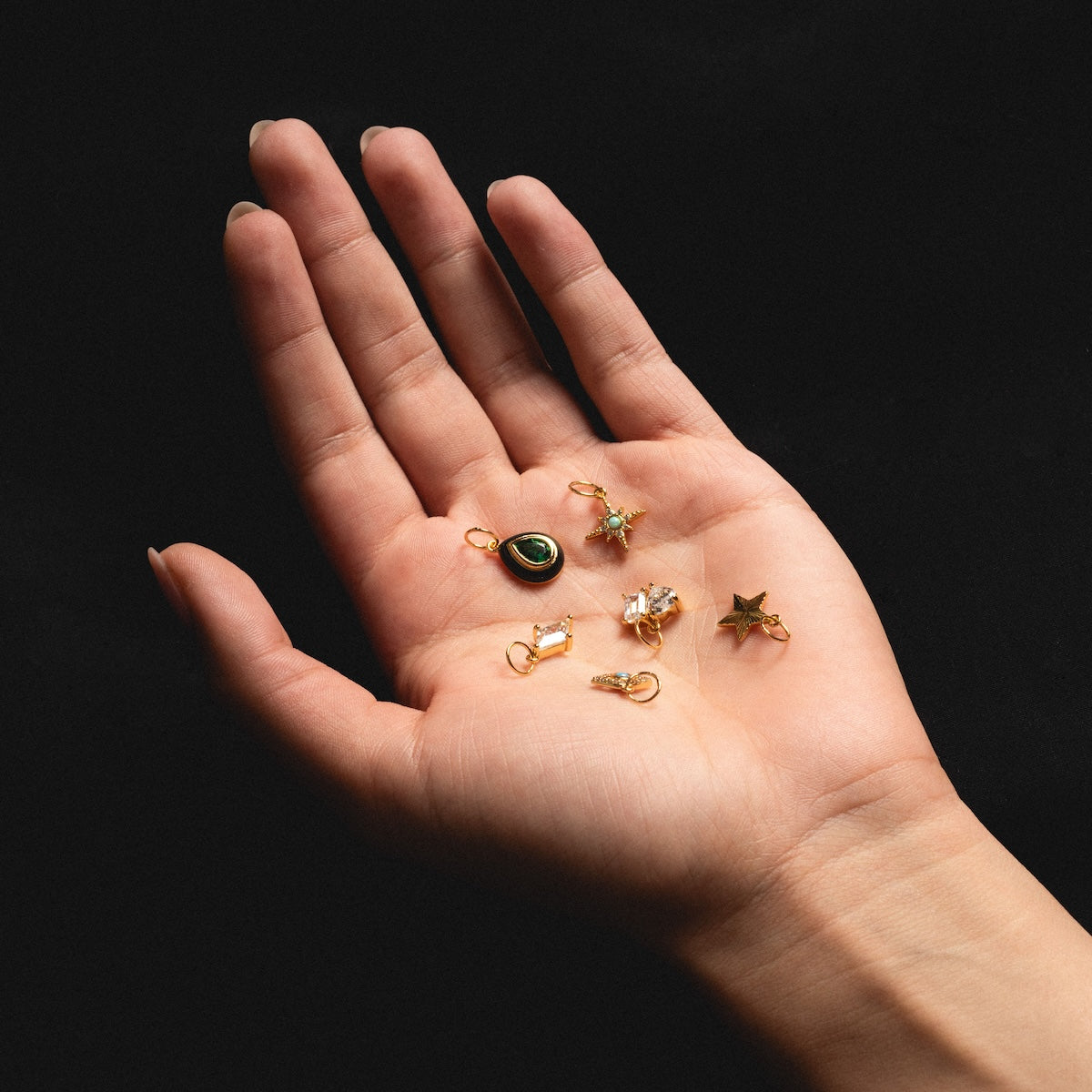 A hand with a neutral background holds five yellow gold plated Charms by Jurate JLA, including a green oval, a cluster of simulated diamonds, a crescent moon, a star, and a round white stone.