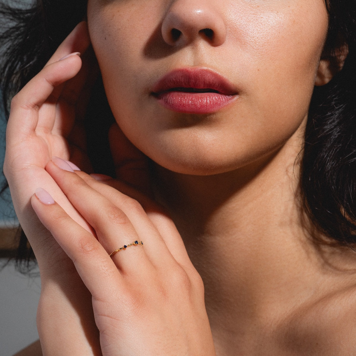 A close-up shows a woman’s manicured hand with natural makeup and pink lips, wearing the Solid Gold 2025 September Capsule Delicate Blue Sapphire Stacking Ring in 9k yellow gold as she gently touches her face.