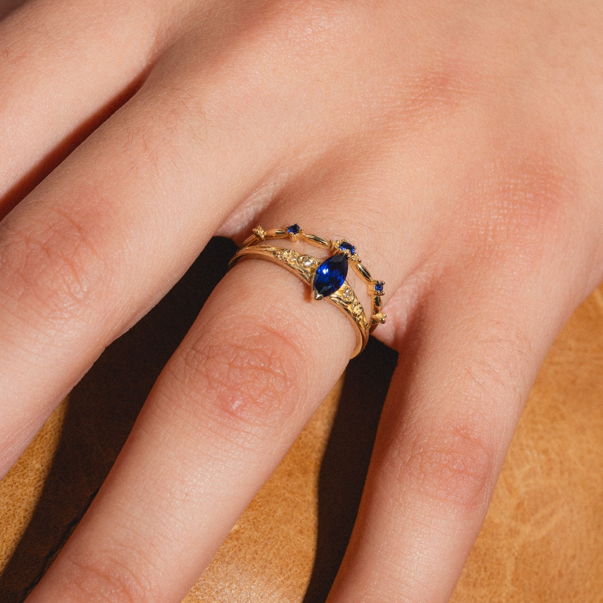 A hand wears the Solid Gold 2025 September Capsule Blue Sapphire Ring Set, featuring a marquise blue sapphire and small blue accents, resting on a brown leather surface. Includes free gift.