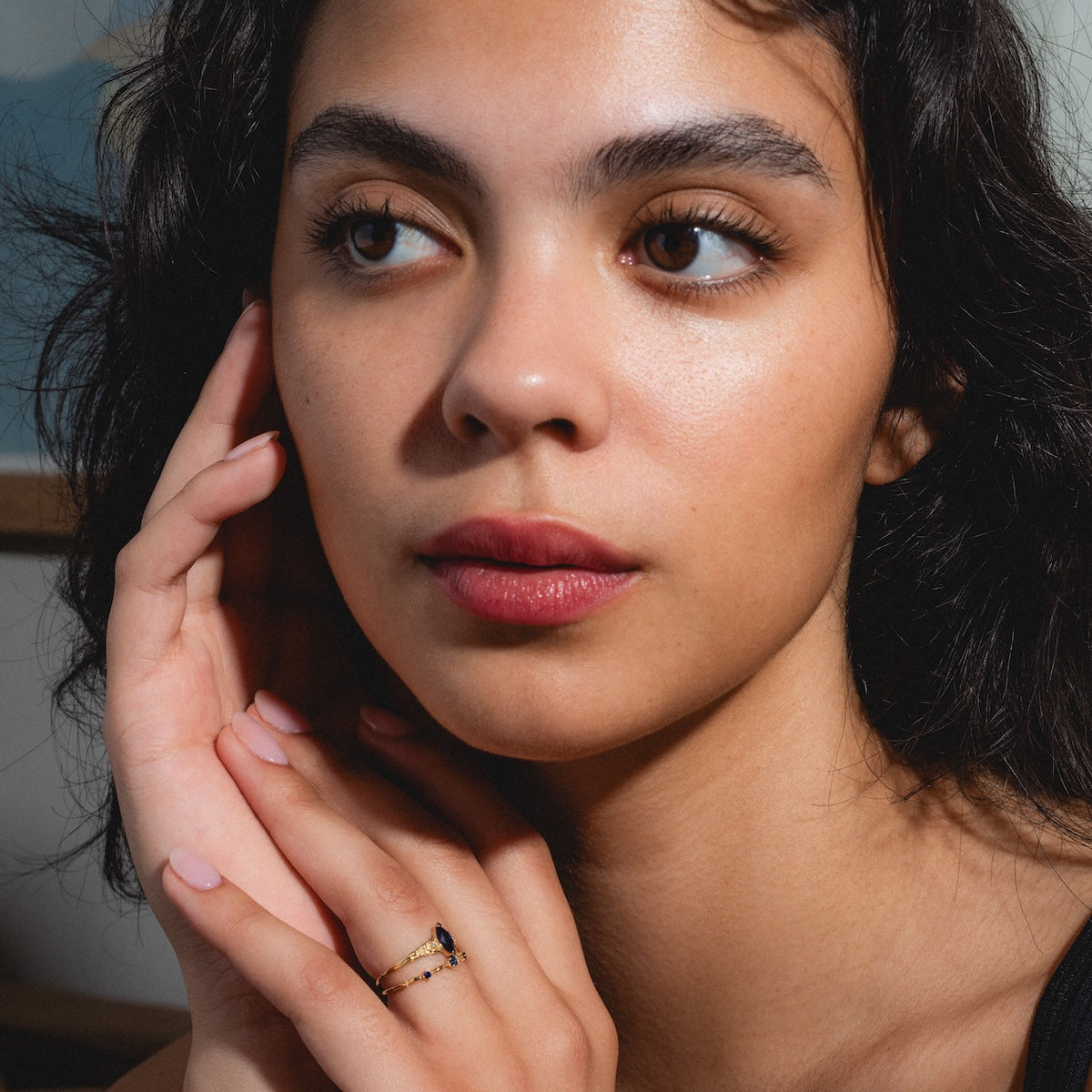 A woman with dark hair gazes thoughtfully to the side, resting her face on her hand. She wears the Solid Gold 2025 September Capsule Blue Sapphire Ring Set with Free Gift and has natural makeup and pink lips.