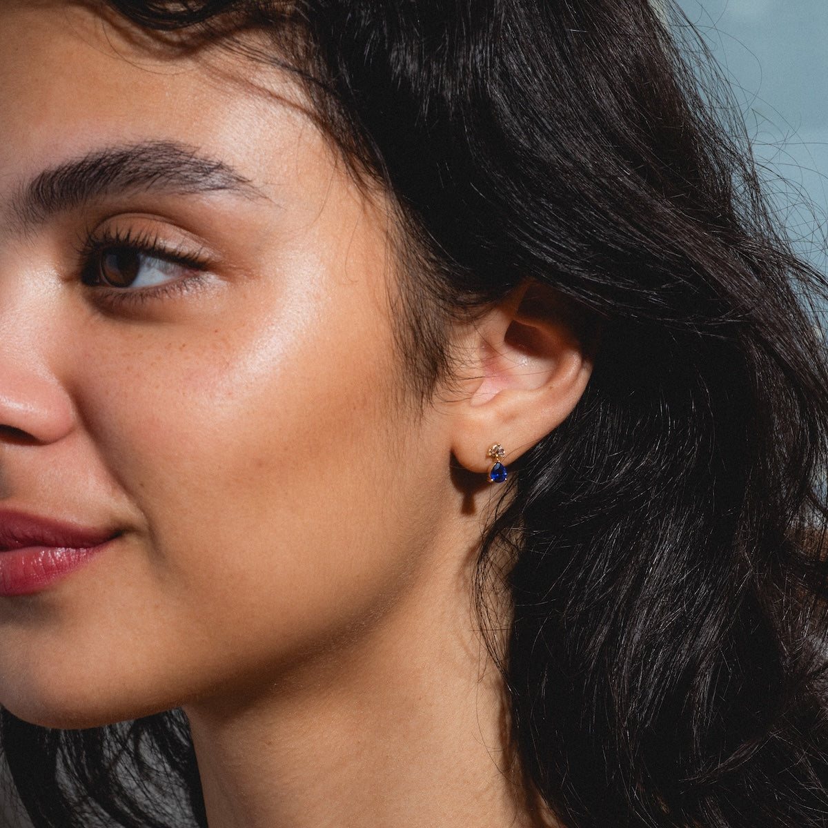 A woman with long dark hair wears Solid Gold 2025 September Capsule Classic Blue Sapphire Stud Earrings, featuring a blue sapphire drop. She has smooth skin, subtle makeup, and gazes softly to the left with a relaxed expression.