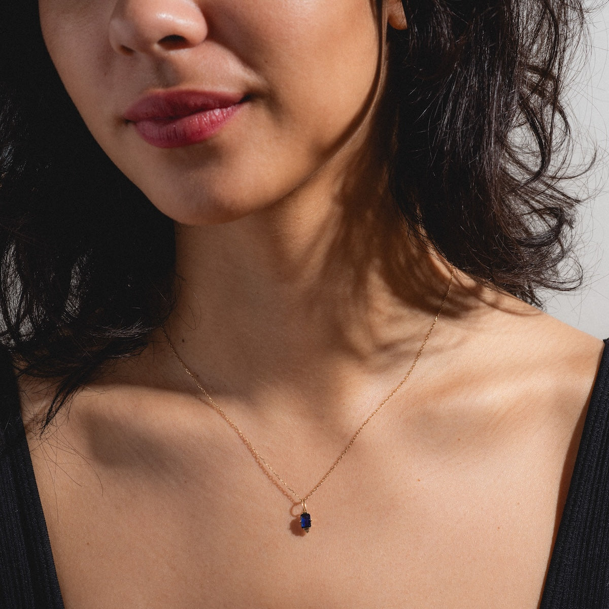 A woman with wavy dark hair wears the Solid Gold 2025 September Capsule Classic Blue Sapphire Pendant Necklace—9k yellow gold with a lab-created blue sapphire. She has natural makeup, soft pink lips, and a black deep neckline top.