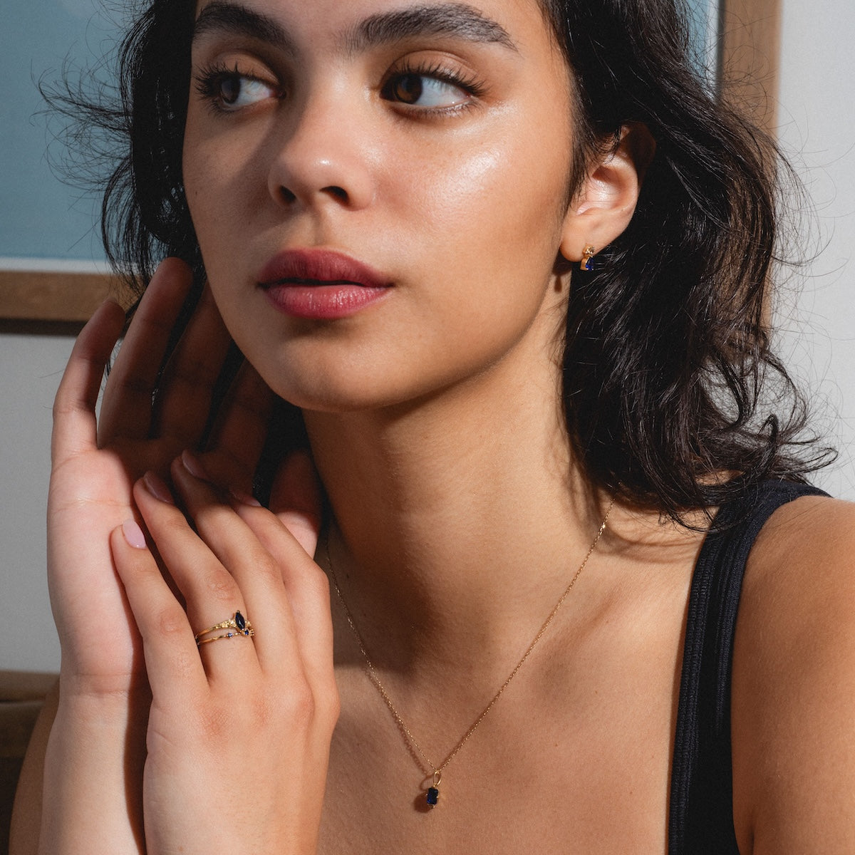 A woman with dark hair and radiant skin wears a black tank, accessorized with the Solid Gold 2025 September Capsule Blue Sapphire Full Set (plus free gift), gazing thoughtfully to the side in a softly-lit indoor space.