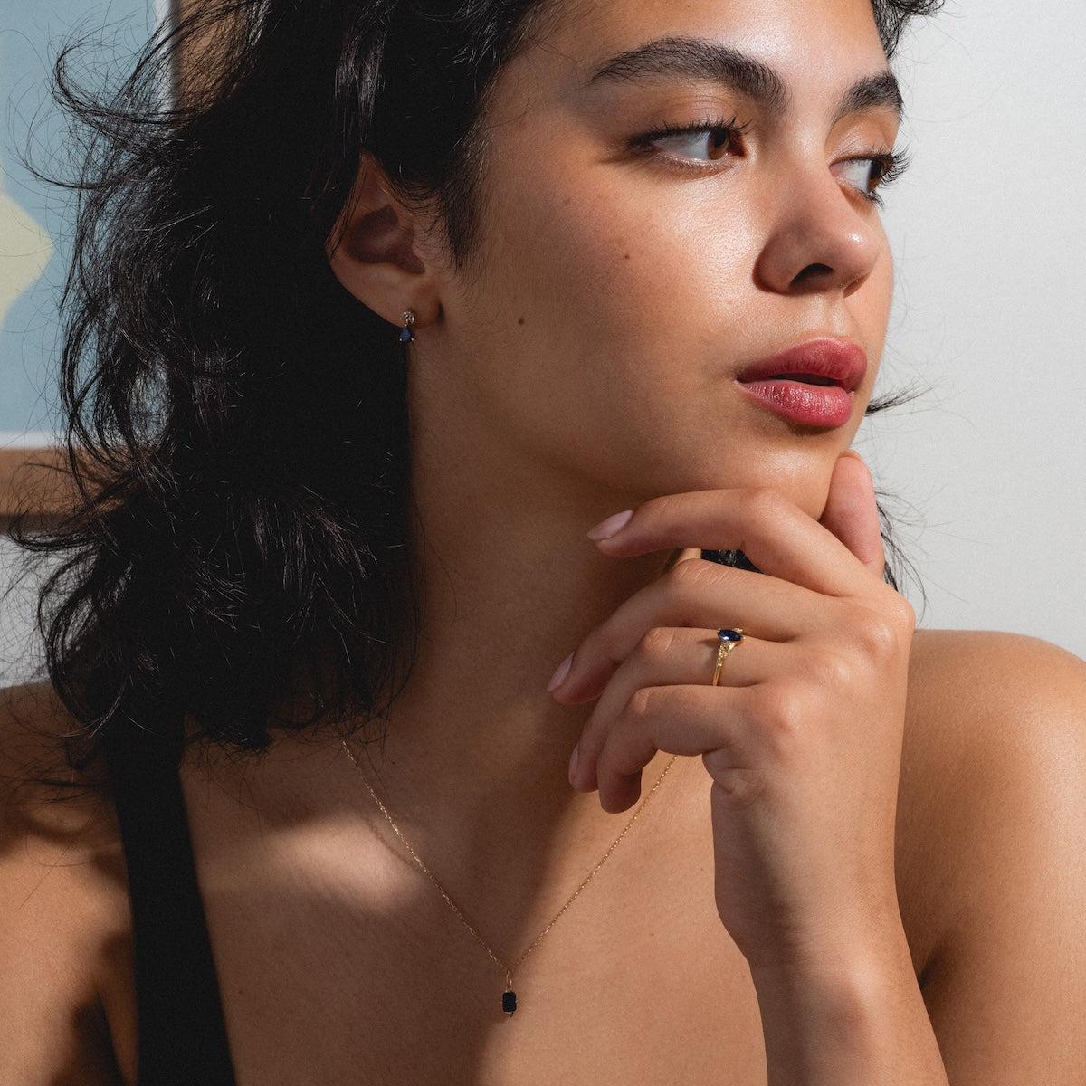 A woman with wavy dark hair gazes to the side, her hand resting on her chin. She wears a black top, simple earrings, and the Solid Gold 2025 September Capsule Blue Sapphire Statement Set with Free Gift—an elegant limited edition look.