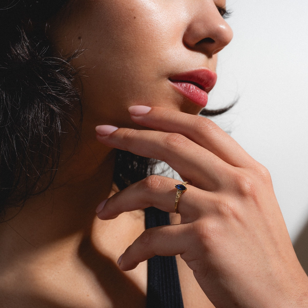 A woman with wavy dark hair and red lipstick touches her face, showcasing the Solid Gold 2025 September Capsule Classic Blue Sapphire Statement Ring on her finger. She wears a black top and has light pink manicured nails.