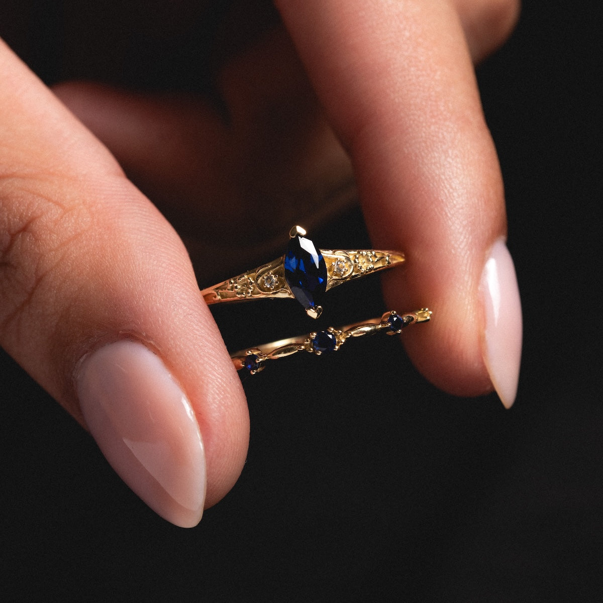 A hand with manicured nails holds the Solid Gold 2025 September Capsule Classic Blue Sapphire Statement Ring—crafted in 9k yellow gold, featuring a marquise-cut lab-created blue sapphire and delicate bands of smaller blue stones.