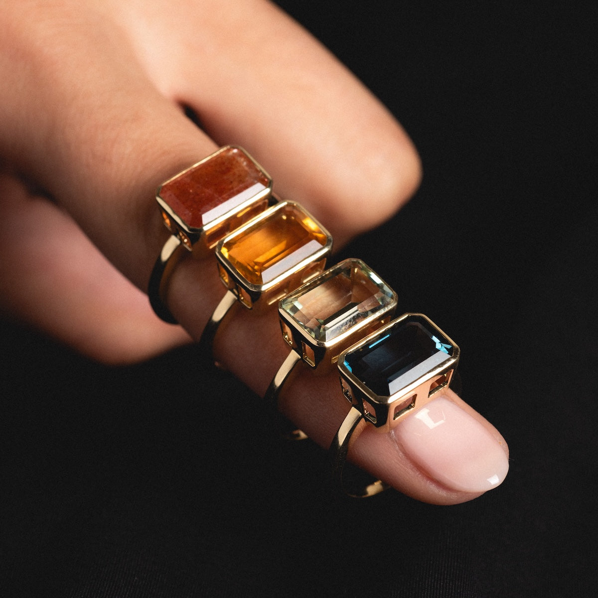 A manicured hand wears four 14k Jojo Rings—solid yellow gold bands, each set with a large rectangular natural gemstone in red, yellow, pale green, and deep blue—modern heirlooms shown against a dark background.