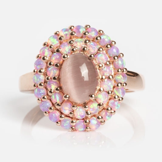 The Rose Gold Pink Opal and Cats Eye Moonstone Crown Ring features a central oval gemstone encircled by two rows of iridescent opals, all set in ornate rose gold, displayed on a plain white background.