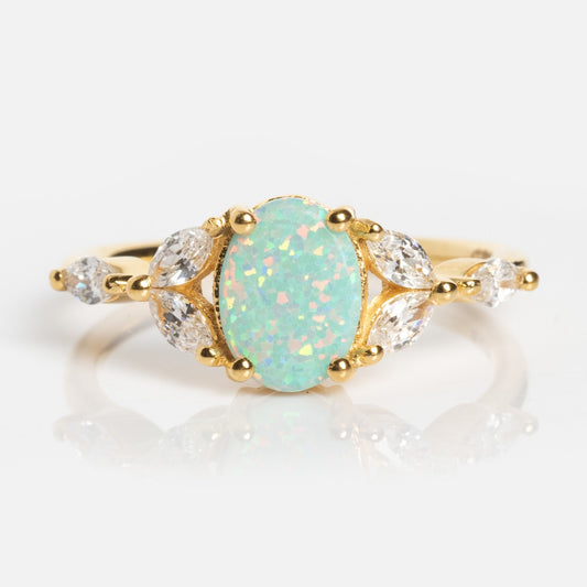 The Opal Enchanted Ring features an oval opal center with iridescent hues, accented by three marquise-cut clear stones on each side, displayed on a white background.