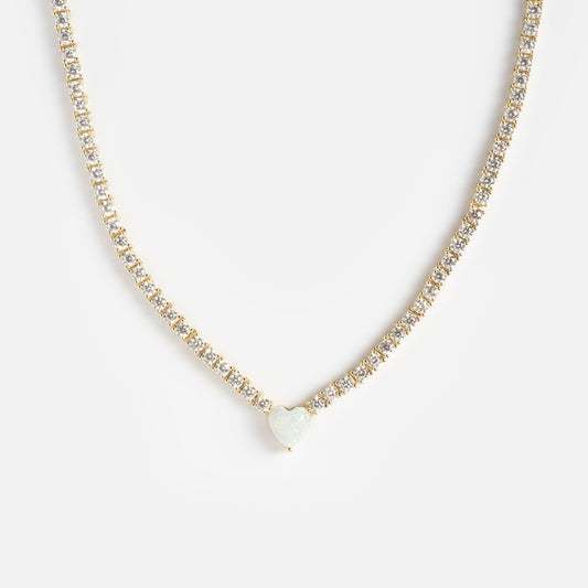 The Queen of Hearts Tennis Necklace is a delicate gold piece featuring small clear gemstones and a heart-shaped opal pendant at the center, shown on a white background.