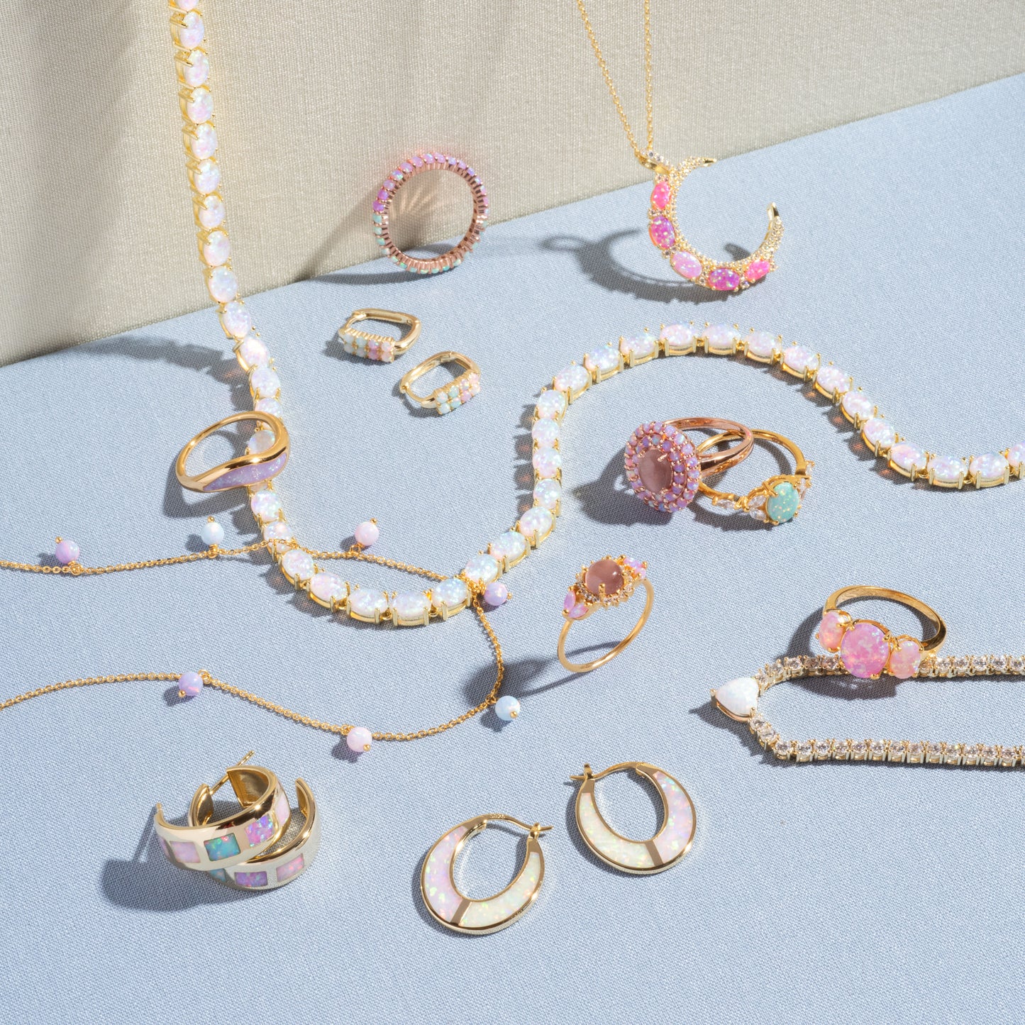 The Opal Dreamers Wave Ring, a gold ring with pink, white, and pastel gemstones, is displayed on light blue and beige fabric backgrounds along with other jewelry pieces.