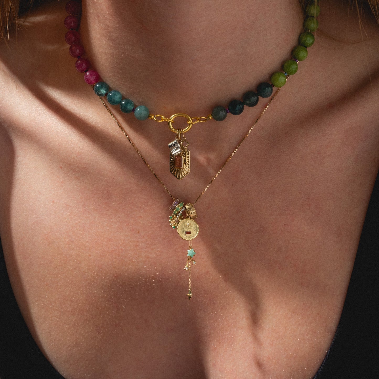 Close-up of a person's neck wearing two necklaces: a beaded green and pink strand featuring an Ethiopian opal and yellow gold charm, and the Solid Gold Lucky Charm necklace with multiple pendants, including a horseshoe, against a black top.