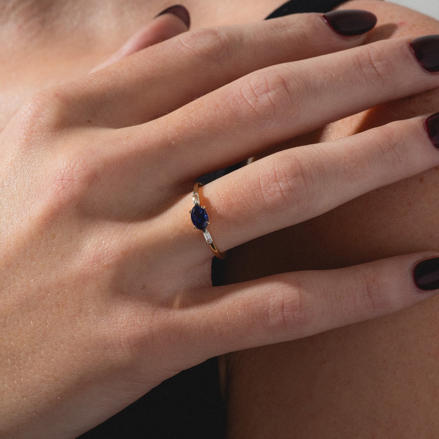A close-up of a hand with dark burgundy nail polish, wearing the Solid Gold Classic Sapphire Ring set with a fissure-filled blue sapphire, resting gently on a shoulder.