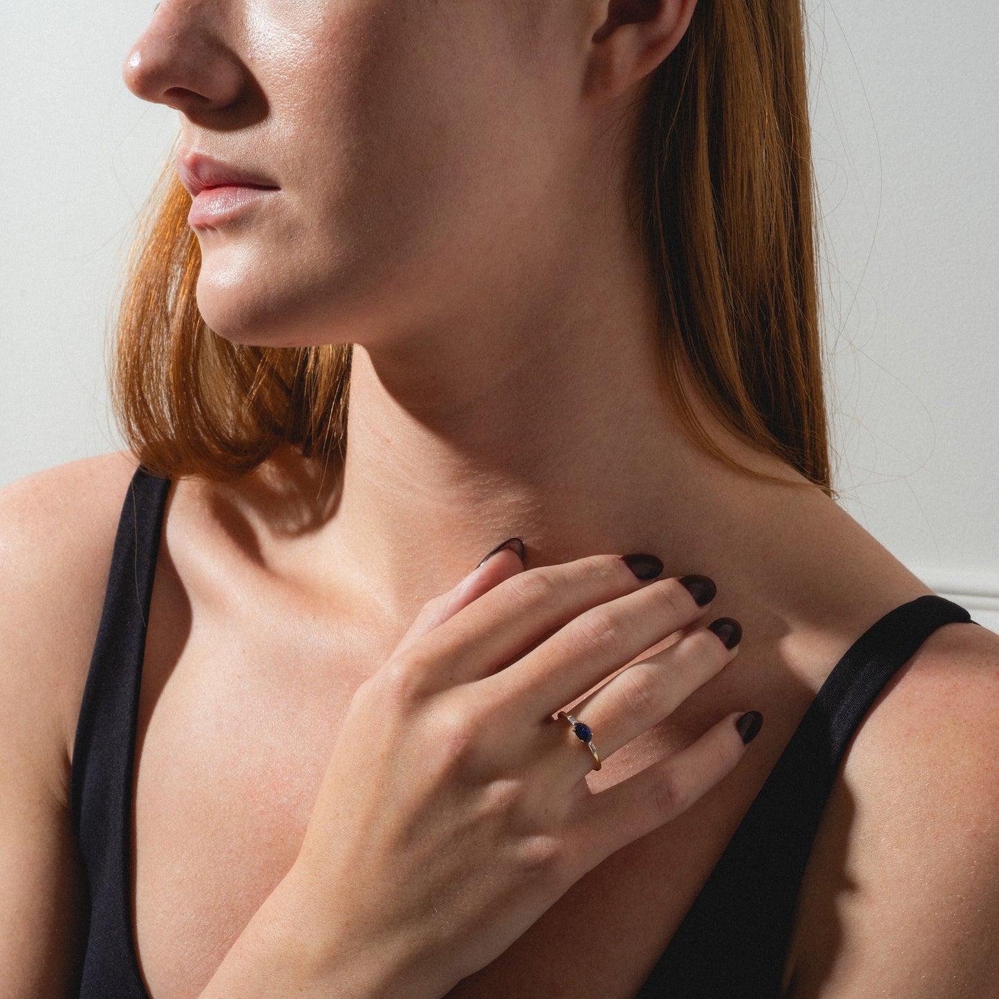A woman with long red hair in a black sleeveless top touches her collarbone, showcasing the Solid Gold Classic Sapphire Ring—crafted from 9k gold and featuring a striking fissure-filled blue sapphire—on her ring finger.