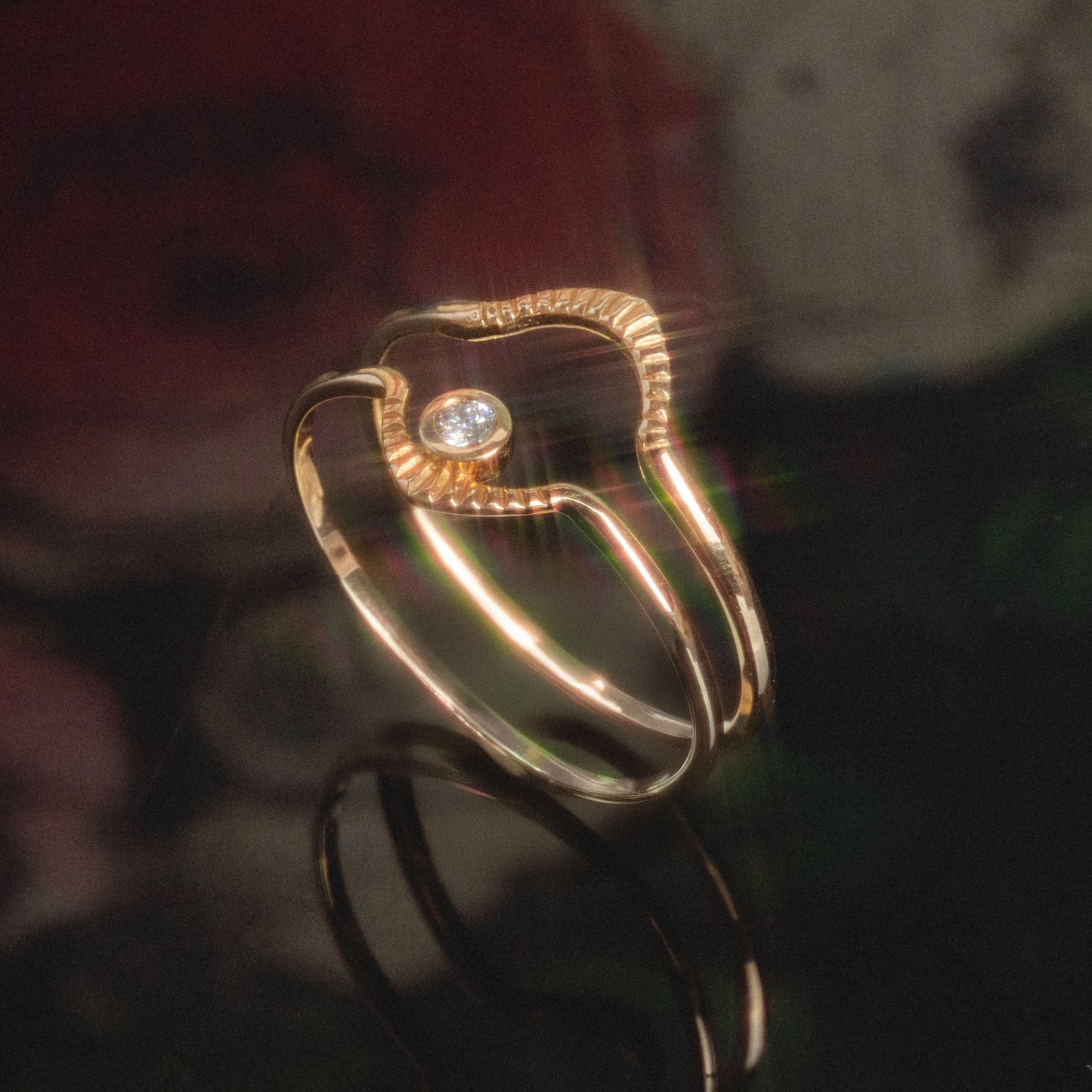 A close-up of the Corrie Ring, featuring a unique wavy gold design with a single round diamond at its center, set against a dark, softly blurred background with hints of red and green.