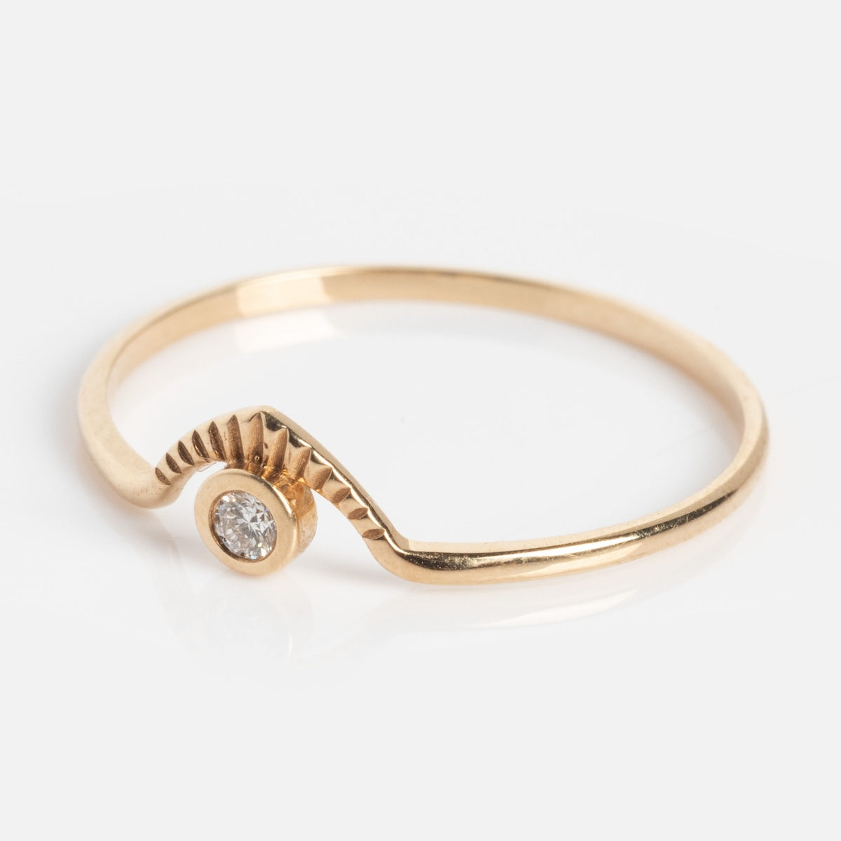 The Corrie Ring is a thin gold band with a small round diamond set off-center in a unique, grooved stepped detail. Its modern, asymmetric shape adds to its contemporary elegance.