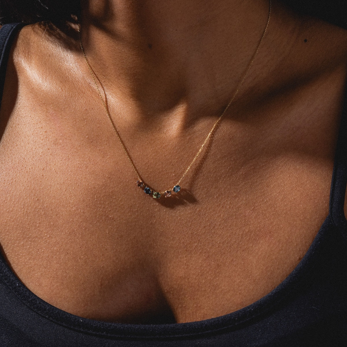 A close-up of a woman’s chest and collarbone shows her wearing the Royal Mile Necklace—a delicate gold chain with small, multicolored gemstones—against her tan skin and a black tank top.