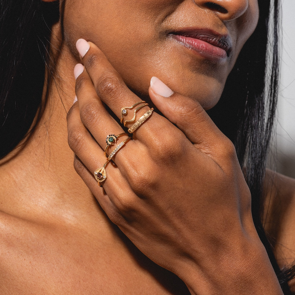 A woman with medium brown skin and long dark hair touches her face, highlighting her neatly manicured pink nails and the Tosia Tourmaline Ring—a solid gold ring featuring a striking bi-color tourmaline—on her finger.