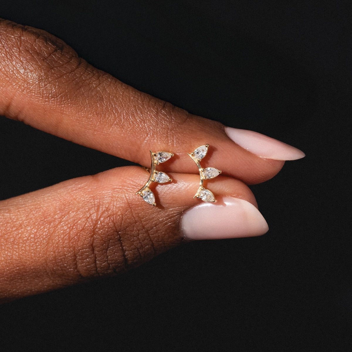 A hand with light pink nails holds the Rain Diamond Studs, gold earrings with three leaf-shaped settings and sparkling clear stones, against a dark background.