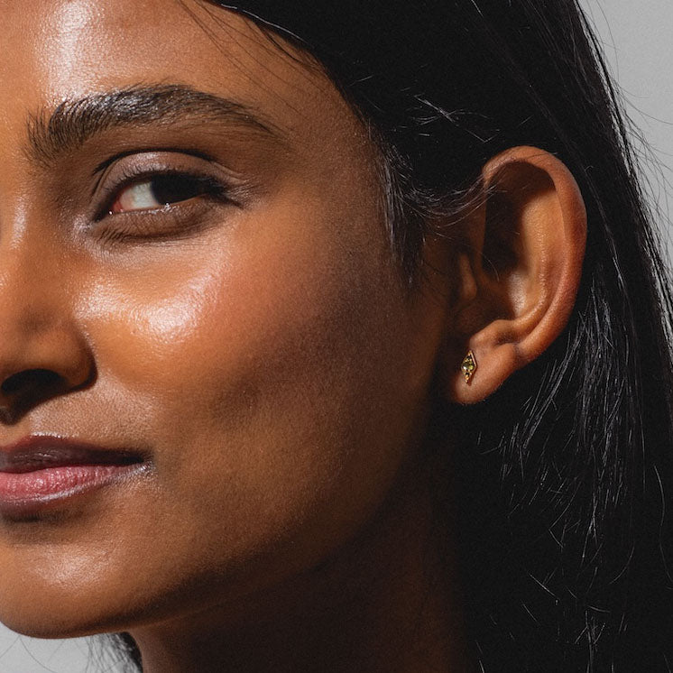 Close-up of a woman in profile with smooth skin, dark hair, and a slight smile, wearing Shillay Black Diamond Studs in 14k yellow gold against a neutral background.